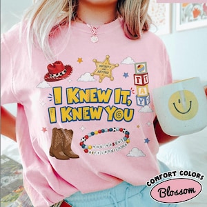 Pode incluir: Uma t-shirt rosa claro com uma estampa gr&aacute;fica que inclui um chap&eacute;u de cowboy vermelho, uma estrela de xerife dourada com a inscri&ccedil;&atilde;o Infinity and Beyond, blocos de letras empilhados, botas de cowboy castanhas e uma pulseira de contas com o texto You've got a friend in me.