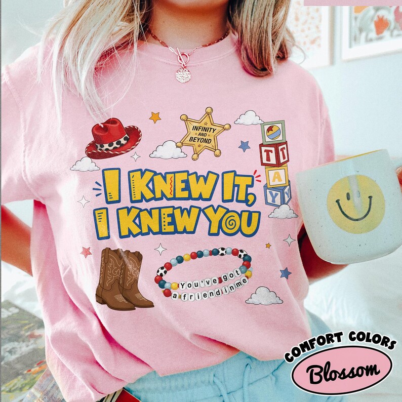 Pode incluir: Uma t-shirt rosa claro com uma estampa gr&aacute;fica que inclui um chap&eacute;u de cowboy vermelho, uma estrela de xerife dourada com a inscri&ccedil;&atilde;o Infinity and Beyond, blocos de letras empilhados, botas de cowboy castanhas e uma pulseira de contas com o texto You've got a friend in me.
