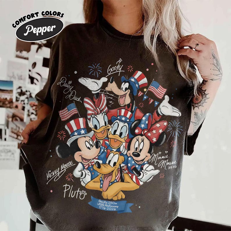 May include: Dark grey t-shirt featuring Mickey Mouse, Minnie Mouse, Donald Duck, Daisy Duck, Goofy, and Pluto in patriotic attire. The shirt commemorates the USA's 250th anniversary, with text and fireworks graphics.