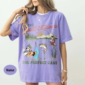 Puede incluir: Camiseta violeta con una escena de pesca de dibujos animados. El dise&ntilde;o incluye el texto "The Great Outdoors" y "The Perfect Cast". El gr&aacute;fico presenta un perro de dibujos animados pescando, un pez y una persona tomando caf&eacute;.