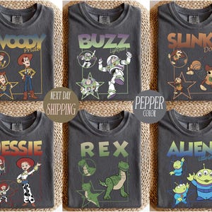 May include: Six charcoal gray t-shirts featuring characters from the animated film Toy Story. Each shirt displays a different character: Woody, Buzz, Slinky Dog, Jessie, Rex, and Aliens. The shirts have character names and images in various colors.