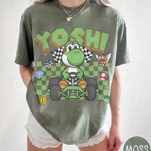 May include: An olive green t-shirt featuring a graphic of the character Yoshi driving a go-kart. The design includes the text YOSHI in large green letters, a checkered flag, a question mark block, a coin, a mushroom, and other game icons.