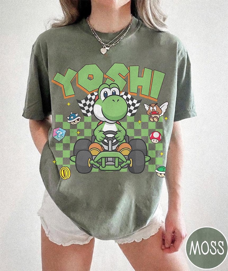May include: An olive green t-shirt featuring a graphic of the character Yoshi driving a go-kart. The design includes the text YOSHI in large green letters, a checkered flag, a question mark block, a coin, a mushroom, and other game icons.