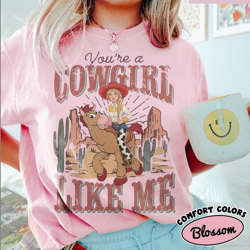 May include: A light pink t-shirt featuring a graphic print of a cartoon cowgirl riding a brown horse in a desert landscape with cacti and rock formations. The text reads You're a COWGIRL LIKE ME in a distressed font. The shirt has a crew neckline.