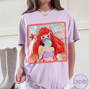 Puede incluir: Una camiseta lila con un gr&aacute;fico colorido de una sirena con pelo rojo, gafas de sol en forma de coraz&oacute;n y una burbuja. El gr&aacute;fico est&aacute; enmarcado con un borde rojo y rodeado de criaturas marinas.