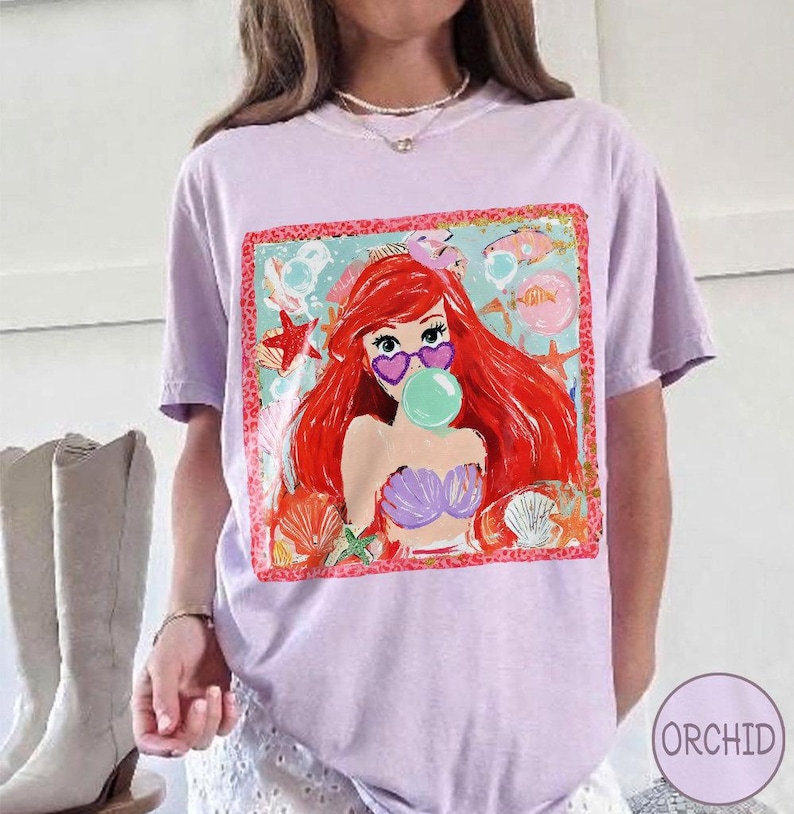 Puede incluir: Una camiseta lila con un gr&aacute;fico colorido de una sirena con pelo rojo, gafas de sol en forma de coraz&oacute;n y una burbuja. El gr&aacute;fico est&aacute; enmarcado con un borde rojo y rodeado de criaturas marinas.