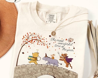 Puede incluir: Una camiseta de color marfil con un gráfico de Winnie the Pooh, Igor, Piglet y Tigger caminando sobre un puente de piedra. El texto dice, The most wonderful time of the year. La camiseta tiene una etiqueta de la marca Comfort Colors.