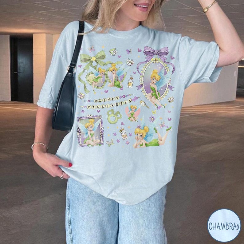 May include: A light blue short-sleeve t-shirt featuring multiple illustrations of Tinkerbell, green bows, and purple frames. The text Disney Tinkerbell is printed in the center. The shirt is paired with blue denim jeans and a black shoulder bag.