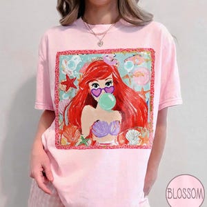 Puede incluir: Camiseta rosa claro con un gr&aacute;fico colorido de una sirena de dibujos animados con cabello rojo, gafas de sol en forma de coraz&oacute;n y una burbuja de chicle. El gr&aacute;fico est&aacute; enmarcado con un borde decorativo y rodeado de conchas marinas y estrellas de mar.