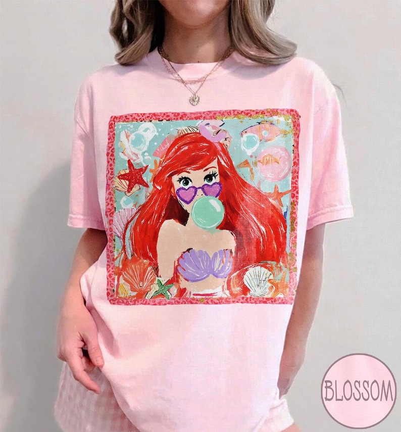 Puede incluir: Camiseta rosa claro con un gr&aacute;fico colorido de una sirena de dibujos animados con cabello rojo, gafas de sol en forma de coraz&oacute;n y una burbuja de chicle. El gr&aacute;fico est&aacute; enmarcado con un borde decorativo y rodeado de conchas marinas y estrellas de mar.