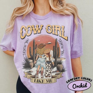 May include: A light purple short-sleeve t-shirt featuring a graphic of two figures in cowboy hats standing in a desert landscape with cacti. The text reads COW GIRL and I KNEW YOU I KNEW YOU LIKE ME. The shirt has a relaxed fit and crew neckline.