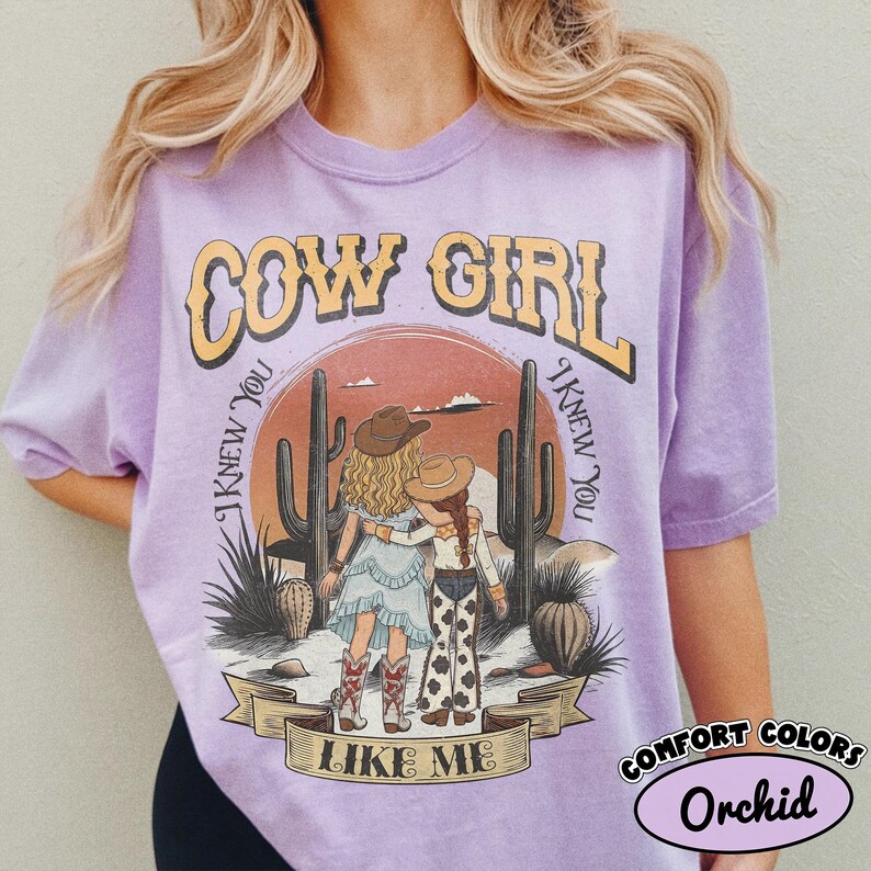 May include: A light purple short-sleeve t-shirt featuring a graphic of two figures in cowboy hats standing in a desert landscape with cacti. The text reads COW GIRL and I KNEW YOU I KNEW YOU LIKE ME. The shirt has a relaxed fit and crew neckline.