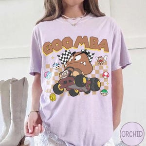 May include: A light purple t-shirt featuring a graphic of a Goomba character driving a go-kart. The design includes a checkered background, the text GOOMBA in brown letters, and various small game icons like mushrooms and shells scattered around.