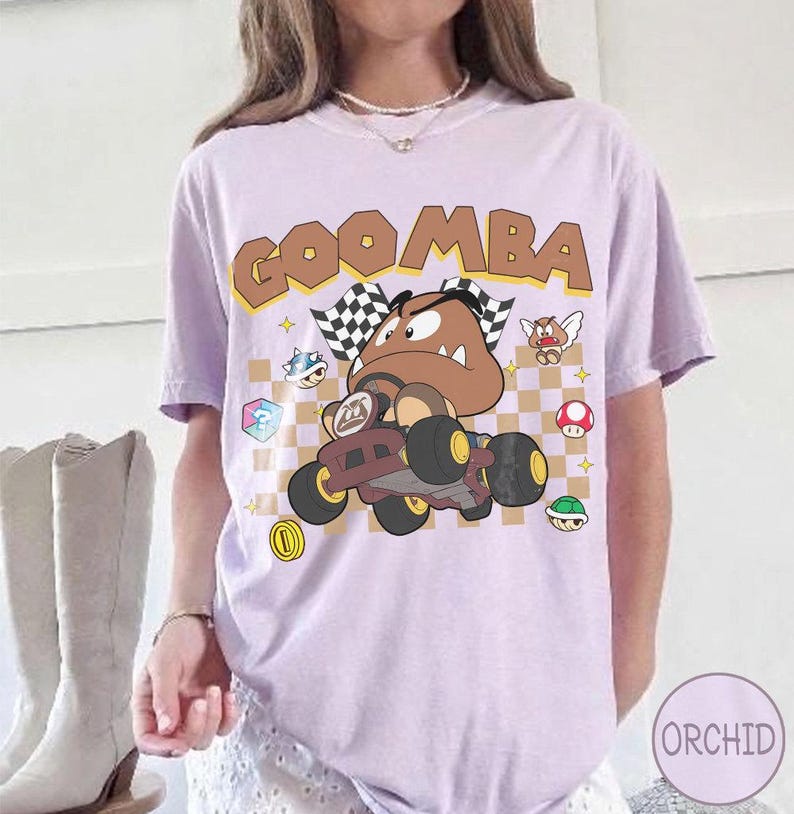 May include: A light purple t-shirt featuring a graphic of a Goomba character driving a go-kart. The design includes a checkered background, the text GOOMBA in brown letters, and various small game icons like mushrooms and shells scattered around.