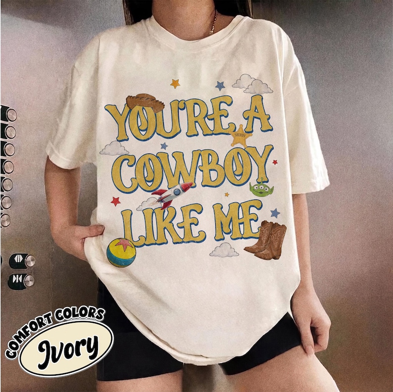 Puede incluir: Una camiseta de color marfil con el texto amarillo "YOU'RE A COWBOY LIKE ME" e ilustraciones gr&aacute;ficas de un sombrero de vaquero marr&oacute;n, un cohete, un alien&iacute;gena verde, una pelota colorida, botas de vaquero marrones, una estrella de sheriff dorada.