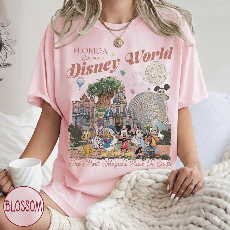 K&ouml;nnte beinhalten: Hellrosa T-Shirt mit einem Disney World-Motiv. Das Design enth&auml;lt den Text "Florida Est. 1971 Disney World" und zeigt Zeichentrickfiguren und Park-Wahrzeichen. Das Shirt hat auch den Satz "The Most Magical Place On Earth."