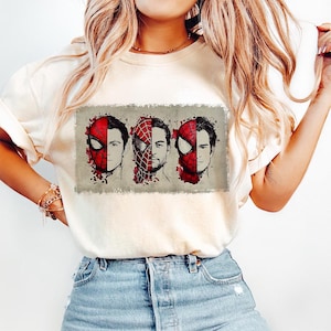 May include: Cream-colored t-shirt featuring a graphic of three Spider-Man faces, each with a different mask design. The faces are set against a rectangular, textured background with red paint splatter details.