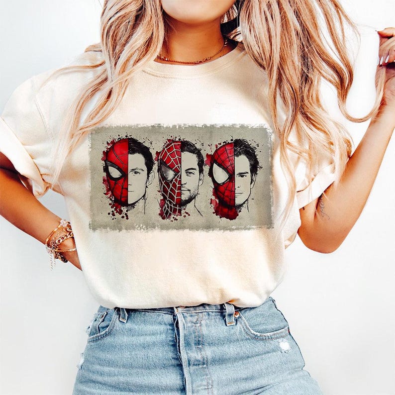 May include: Cream-colored t-shirt featuring a graphic of three Spider-Man faces, each with a different mask design. The faces are set against a rectangular, textured background with red paint splatter details.