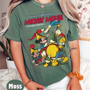 May include: Moss green t-shirt featuring a graphic of Mickey Mouse and friends in a band concert scene. The text "Walt Disney's Mickey Mouse" is displayed above the image. The shirt has short sleeves and a crew neck.