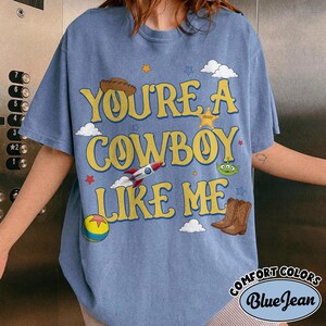 Puede incluir: Una camiseta azul con el texto amarillo "YOU'RE A COWBOY LIKE ME" rodeado de gr&aacute;ficos que incluyen un sombrero de vaquero marr&oacute;n, una estrella de sheriff, un cohete, un alien&iacute;gena verde de tres ojos, una pelota colorida y nubes blancas.