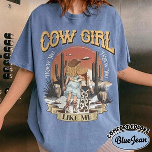 May include: A blue short-sleeve t-shirt featuring a graphic of two figures in cowboy hats standing in a desert landscape with cacti. The text reads COW GIRL and I KNEW YOU I KNEW YOU LIKE ME. The shirt has a crew neckline and a relaxed fit.