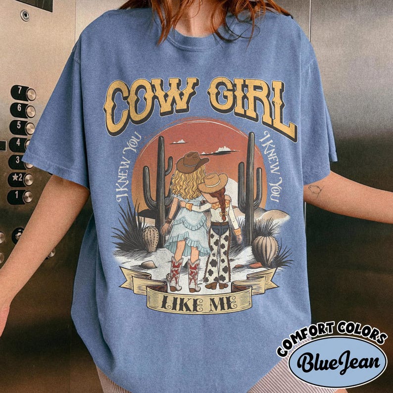 May include: A blue short-sleeve t-shirt featuring a graphic of two figures in cowboy hats standing in a desert landscape with cacti. The text reads COW GIRL and I KNEW YOU I KNEW YOU LIKE ME. The shirt has a crew neckline and a relaxed fit.