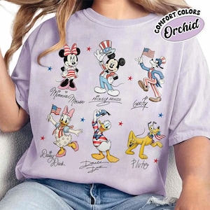 May include: Orchid-coloured t-shirt featuring cartoon characters Minnie Mouse, Mickey Mouse, Goofy, Daisy Duck, Donald Duck, and Pluto in patriotic outfits. The shirt has the words "COMFORT COLORS" and "Orchid" printed on it.