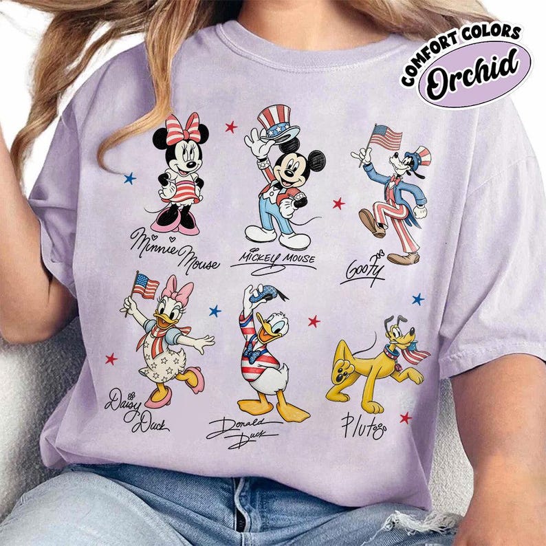 May include: Orchid-coloured t-shirt featuring cartoon characters Minnie Mouse, Mickey Mouse, Goofy, Daisy Duck, Donald Duck, and Pluto in patriotic outfits. The shirt has the words "COMFORT COLORS" and "Orchid" printed on it.