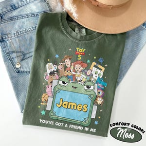 May include: An olive green t-shirt featuring a Toy Story 5 graphic with various characters surrounding a central green frog-shaped frame containing the name James, with the text You've got a friend in me printed below the graphic.