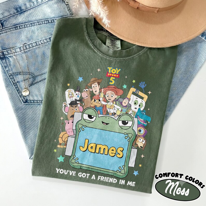 May include: An olive green t-shirt featuring a Toy Story 5 graphic with various characters surrounding a central green frog-shaped frame containing the name James, with the text You've got a friend in me printed below the graphic.