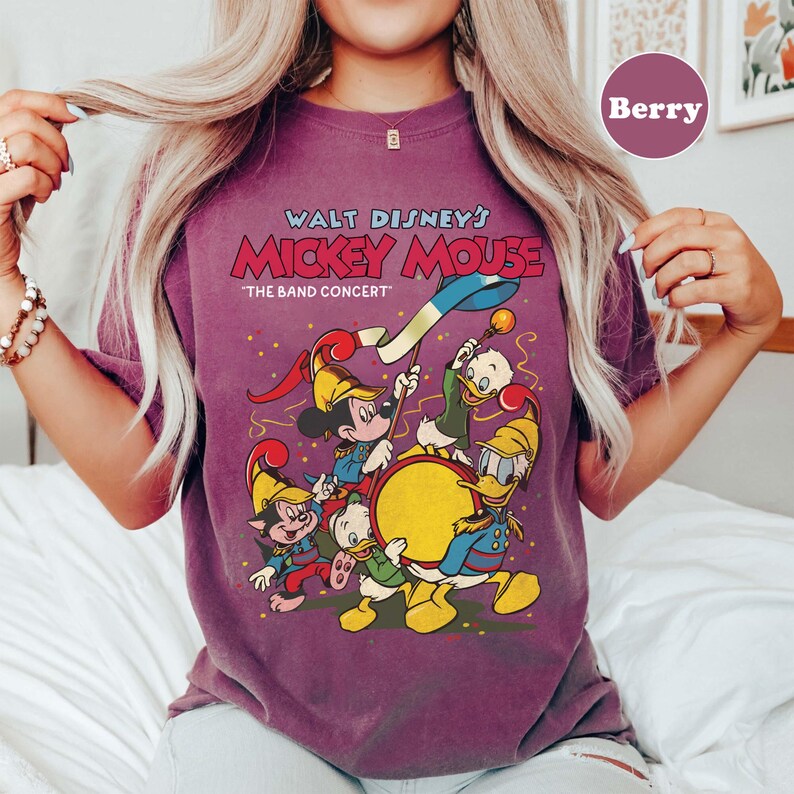 May include: Berry-colored t-shirt featuring a vintage-style graphic of Mickey Mouse and friends in a band concert. The text "Walt Disney's Mickey Mouse" and "The Band Concert" are displayed above the characters. The design includes Mickey, Donald, and other Disney characters.