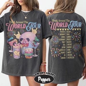May include: A charcoal grey t-shirt featuring a graphic print on the front and back. The front displays a disco ball, cocktails, and the text "Drink Around The World Tour." The back lists countries and toasts with fireworks and mouse ear icons.