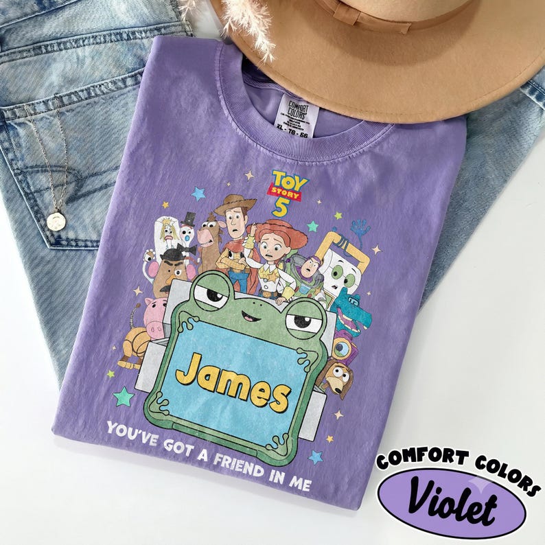 May include: A violet t-shirt featuring a graphic print of Toy Story 5 characters surrounding a green frog-shaped frame with the name James printed in yellow. The text You've Got A Friend In Me is printed below the frame. The shirt has a Comfort Colors label.