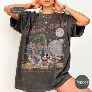 K&ouml;nnte beinhalten: Dunkelgraues T-Shirt mit einem farbenfrohen Disney World-Grafikdruck, der Charaktere und Wahrzeichen zeigt. Der Text "FLORIDA Disney World" und "The Most Magical Place On Earth" ist aufgedruckt. Das Shirt hat einen Vintage-Look.