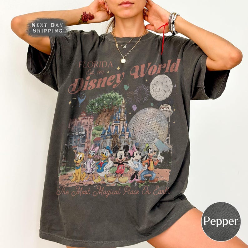 K&ouml;nnte beinhalten: Dunkelgraues T-Shirt mit einem farbenfrohen Disney World-Grafikdruck, der Charaktere und Wahrzeichen zeigt. Der Text "FLORIDA Disney World" und "The Most Magical Place On Earth" ist aufgedruckt. Das Shirt hat einen Vintage-Look.