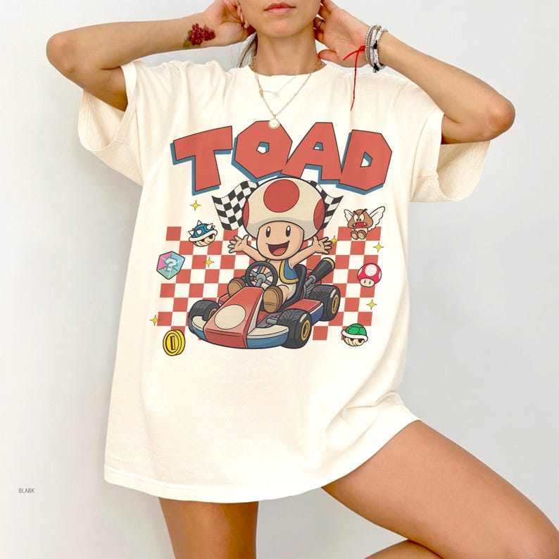 May include: An off-white short-sleeve t-shirt featuring a graphic of the character Toad driving a red go-kart. The design includes the word TOAD in large red letters, a checkered flag, a red and white checkered background, and various game icons.