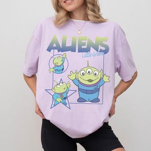 May include: A light purple t-shirt featuring the text "ALIENS Little Green" in a blue and green gradient. The shirt has three cartoon green aliens in blue outfits. One alien is inside a blue star, and another is inside a blue rectangle.