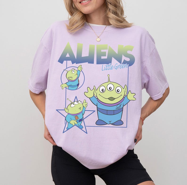 May include: A light purple t-shirt featuring the text "ALIENS Little Green" in a blue and green gradient. The shirt has three cartoon green aliens in blue outfits. One alien is inside a blue star, and another is inside a blue rectangle.