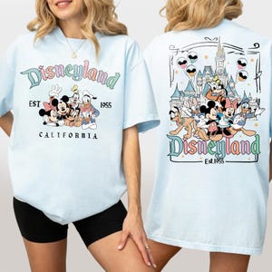 Może przedstawiać: Jasnoniebieska koszulka z logo Disneyland i postaciami z kresk&oacute;wek, takimi jak Myszka Miki i Myszka Minnie, Goofy i Kaczor Donald. Tył koszulki przedstawia zamek i więcej postaci. Widoczny jest r&oacute;wnież tekst "EST 1955" i "CALIFORNIA".