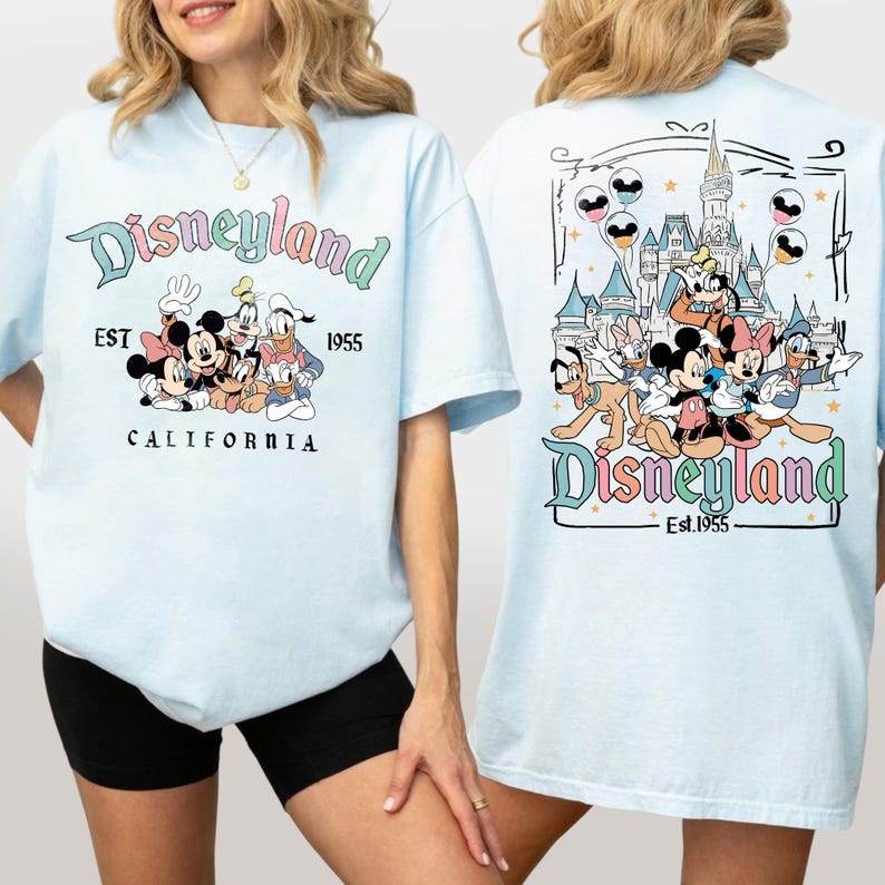 Może przedstawiać: Jasnoniebieska koszulka z logo Disneyland i postaciami z kresk&oacute;wek, takimi jak Myszka Miki i Myszka Minnie, Goofy i Kaczor Donald. Tył koszulki przedstawia zamek i więcej postaci. Widoczny jest r&oacute;wnież tekst "EST 1955" i "CALIFORNIA".