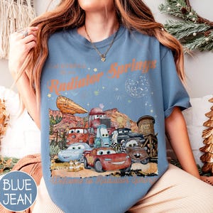 May include: Blue jean t-shirt featuring a graphic of characters from the movie Cars, with the text "California Route 66 Radiator Springs" and "Welcome to Radiator Springs". The design includes cartoon cars and desert scenery.