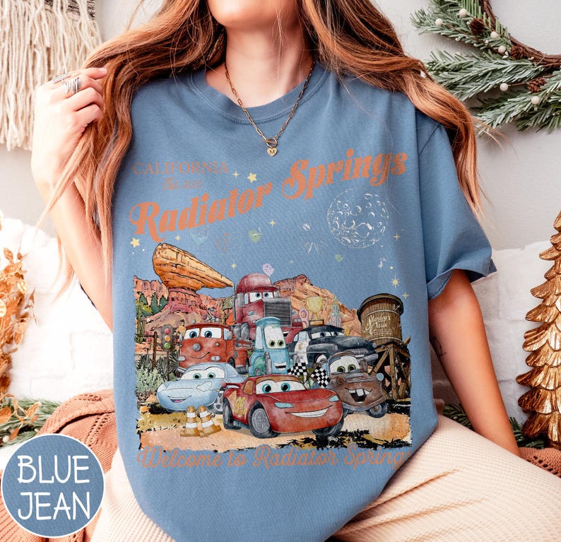 May include: Blue jean t-shirt featuring a graphic of characters from the movie Cars, with the text "California Route 66 Radiator Springs" and "Welcome to Radiator Springs". The design includes cartoon cars and desert scenery.