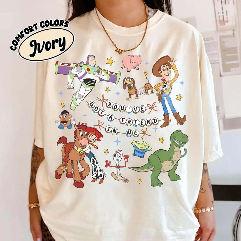 May include: Ivory-coloured t-shirt with a graphic of animated film characters. The design includes Buzz Lightyear, Woody, and other characters, with the phrase "YOU'VE GOT A FRIEND IN ME".