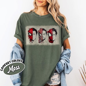 May include: Moss green t-shirt featuring a graphic of three Spider-Man faces, each partially masked in red. The design is printed on a Comfort Colors tee. The shirt is worn with a denim jacket.