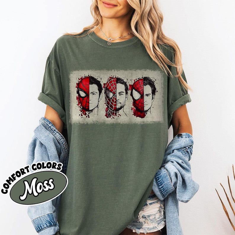 May include: Moss green t-shirt featuring a graphic of three Spider-Man faces, each partially masked in red. The design is printed on a Comfort Colors tee. The shirt is worn with a denim jacket.