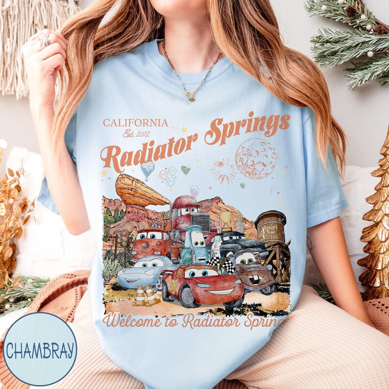 May include: Light blue t-shirt featuring a colorful graphic of the town of Radiator Springs with the text "California Est. 2012 Radiator Springs" and "Welcome to Radiator Springs". The graphic includes various cartoon cars and desert scenery.