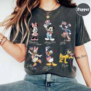 May include: Dark grey t-shirt featuring cartoon characters Minnie Mouse, Mickey Mouse, Goofy, Daisy Duck, Donald Duck, and Pluto in patriotic outfits. Each character is labelled with their name in cursive script. Red and blue stars are scattered around the characters.