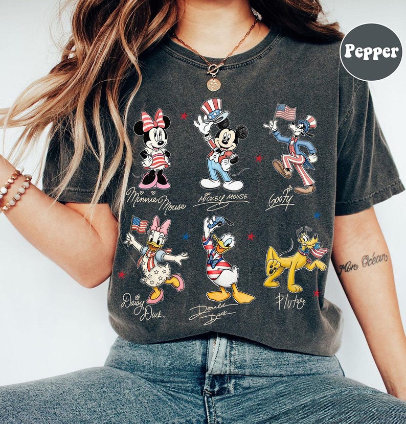 May include: Dark grey t-shirt featuring cartoon characters Minnie Mouse, Mickey Mouse, Goofy, Daisy Duck, Donald Duck, and Pluto in patriotic outfits. Each character is labelled with their name in cursive script. Red and blue stars are scattered around the characters.