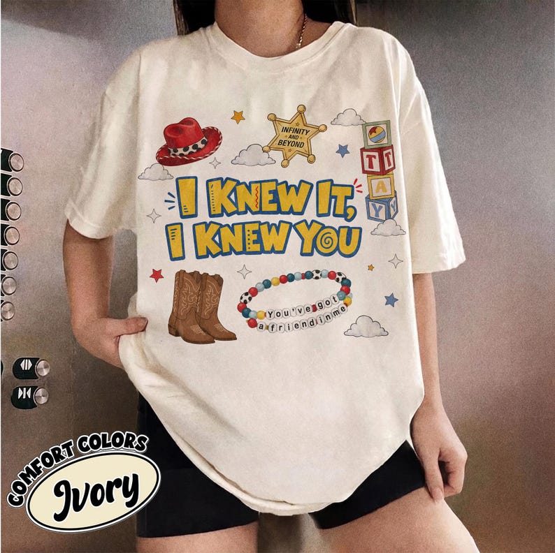 Pode incluir: Uma t-shirt cor de marfim com uma impress&atilde;o gr&aacute;fica com o texto I KNEW IT, I KNEW YOU. O design inclui um chap&eacute;u de cowboy vermelho, uma estrela de xerife dourada, blocos de alfabeto, botas de cowboy castanhas e uma pulseira de contas com o texto You've got a friend in me.
