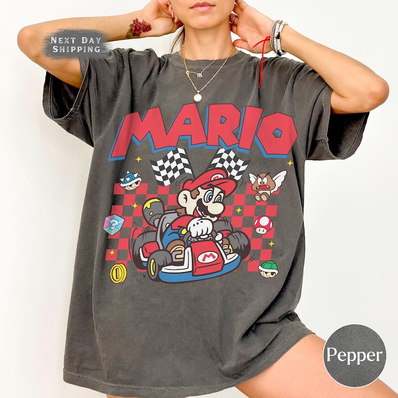 May include: A charcoal gray short-sleeve t-shirt featuring a graphic of Mario driving a go-kart. The design includes the word MARIO in large red letters, a red and black checkered background, and various game icons like a shell and a mushroom.